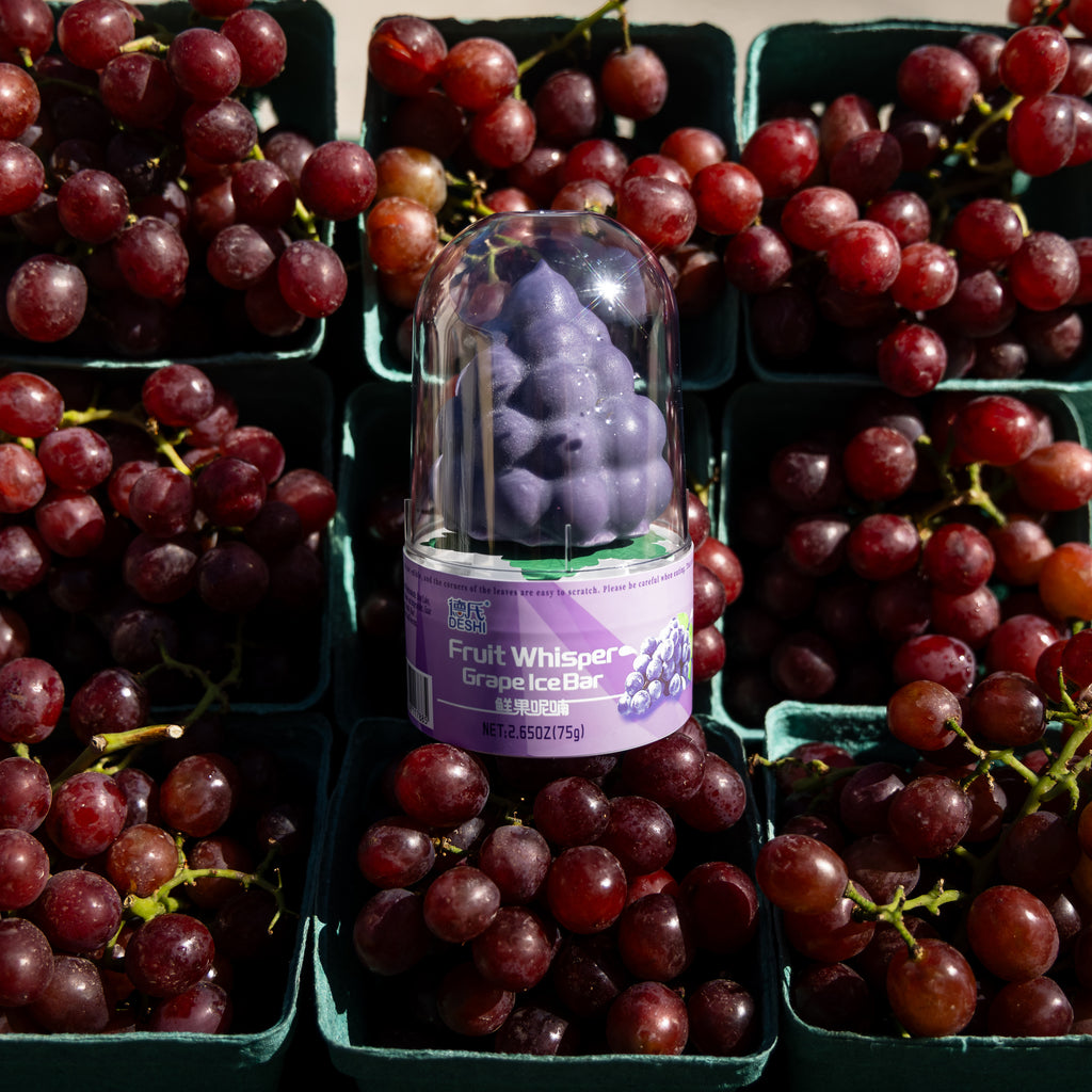 Whole Deshi grape ice pop in its clear dome, surrounded by baskets of grapes