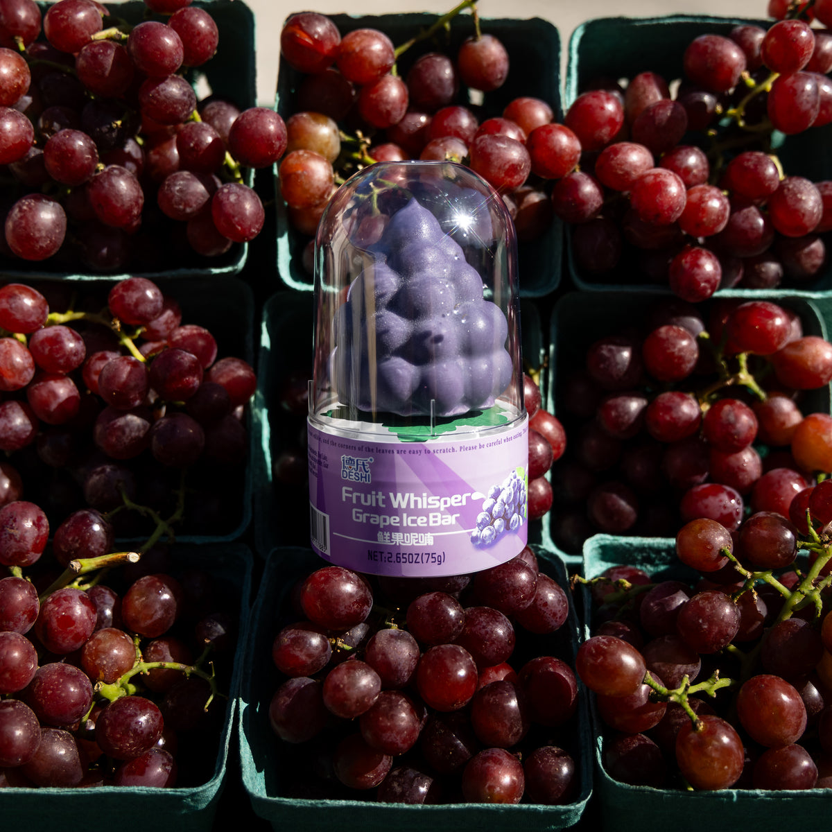 Whole Deshi grape ice pop in its clear dome, surrounded by baskets of grapes